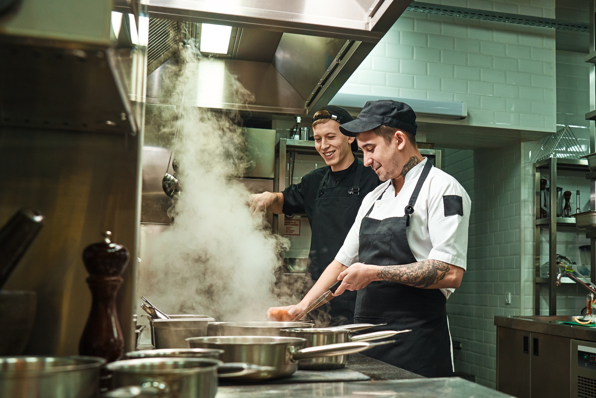 Positive mood. Side view of two cheerful and smiling cooks in uniform are preparing a food in a